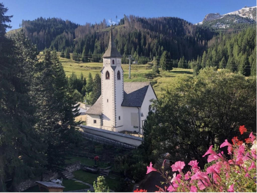 A church with a steeple in the middle of a valley surrounded by greenery in the Dolomites.